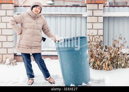 Una donna asiatica di mezza età con cappotto e cappello beige spinge un grande cestino blu nella neve in un ambiente invernale Foto Stock