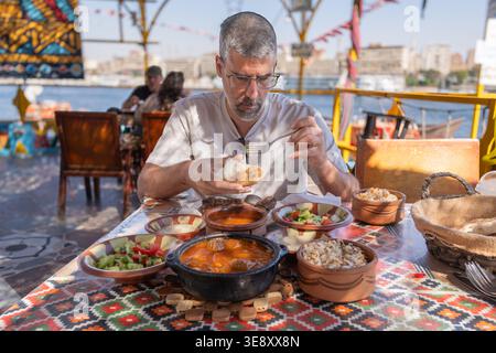 Uomo di mezza età che gusta un pasto egiziano tradizionale in una terrazza all'aperto vicino al fiume Nilo. Concetto di viaggio e cultura alimentare. Foto Stock