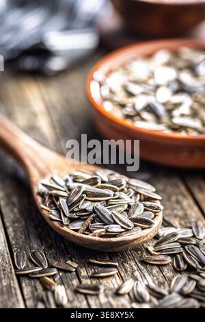 Primo piano di semi di girasole a strisce, ammassati in un cucchiaio di legno con una ciotola di terracotta su un tavolo di legno intemprato, evocando cucina rustica, snack salutari, Foto Stock