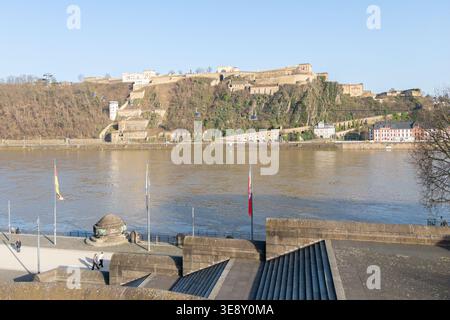 Coblenza, Germania - 02.26.2026: Vista dal Deutsches Eck di Coblenza alla Fortezza di Ehrenbreitstein e al Reno con bandiere cielo azzurro Foto Stock