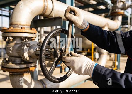 Primo piano di una mano con i guanti che gira una grande valvola industriale, controllando il flusso dei fluidi in un impianto Foto Stock