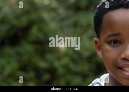 Bambino afroamericano che sorride, guarda la macchina fotografica nel parco con cappuccio grigio, copia spazio. Ritratto, esterno, natura, vegetazione, bokeh, illuminazione, occhio Foto Stock