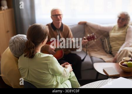 Uomo anziano che suona la chitarra per i residenti anziani e infermiera femminile in casa di cura, sostenendo l'impegno sociale e la cura degli anziani Foto Stock