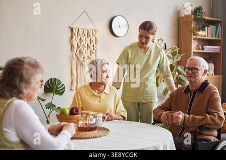 Infermiere senior che assiste i residenti anziani durante il tè nella casa di cura, sostenendo l'assistenza quotidiana e l'interazione sociale Foto Stock