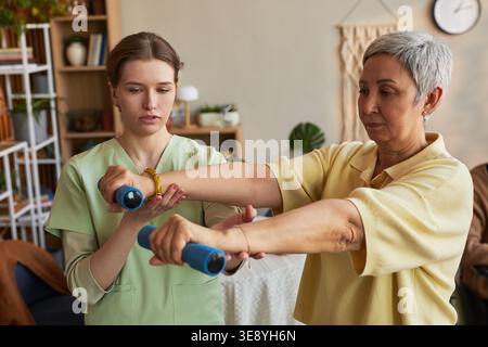 Donna anziana che solleva i manubri durante la riabilitazione guidata con una giovane infermiera in casa di cura Foto Stock
