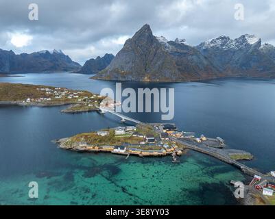 Vista aerea dell'isola di Sakrisoya con le sue iconiche cabine gialle rorbu e un ponte sulle acque turchesi sullo sfondo di montagne innevate in M Foto Stock