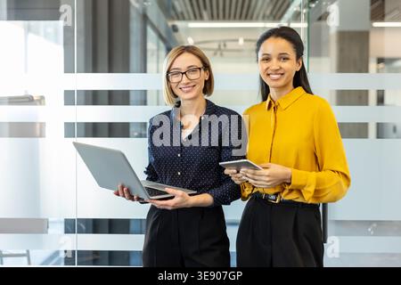 Due donne d'affari multietniche sorridenti in un ufficio luminoso, una con un notebook e l'altra con un tablet digitale, che rappresentano il lavoro di squadra e la collaborazione professionale Foto Stock