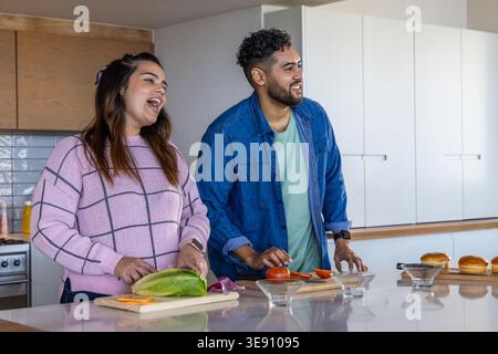 Coppia ridendo e affettando romaine e pomodoro su un'isola di cucina in marmo con taglieri e ciotole. Duo, cucina casalinga, cucina, luce del sole, nazionale, buon appetito Foto Stock