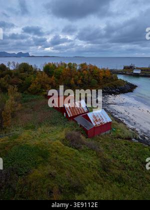 Vista aerea delle tradizionali cabine in legno rosso annidate tra il fogliame autunnale sulla costa rocciosa sotto un cielo nuvoloso a Senja, Troms, Norvegia. Foto Stock