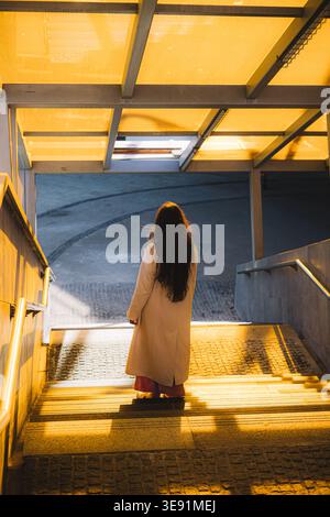 Una giovane donna con un cappotto beige che cammina giù per le scale della città al tramonto Foto Stock