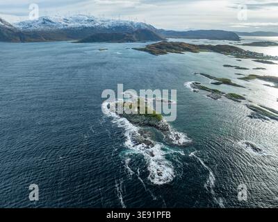 Veduta aerea di una piccola isola rocciosa con un faro bianco circondato dalle acque blu del Mar di Norvegia e dalle montagne innevate del Sommaroy, Foto Stock