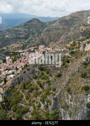 Vista aerea del tortuoso sentiero in pietra sulla ripida scogliera che si affaccia sui tetti storici e sull'aspro paesaggio montano di Taormina, Sicilia, Ital Foto Stock