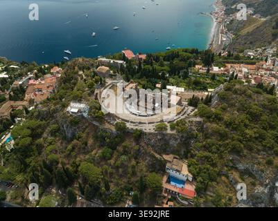 Vista aerea dell'Antico Teatro di Taormina con le sue storiche rovine in pietra che si affacciano sul blu del Mar Ionio e sulle lussureggianti colline verdi di Taormina, Sicilia, Foto Stock