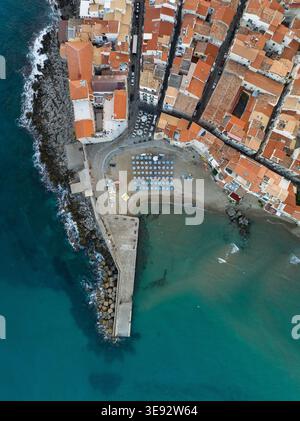 Veduta aerea di Cefalù con tetti arancioni, un molo in pietra e una spiaggia sabbiosa con ombrelloni blu lungo il mare turchese di Cefalù, Sicilia, Italia. Foto Stock