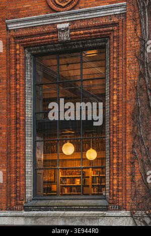 Finestra in mattoni rossi con vista su un'accogliente biblioteca, file di libri sugli scaffali, luci a guanti a pendente calde, architettura storica, atmosfera accademica Foto Stock