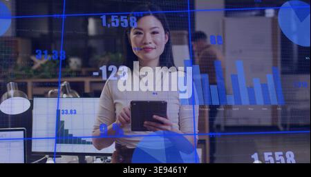 Donna asiatica in piedi che tiene un tablet, indossa la parte superiore beige in un ufficio aperto e mostra sovrapposizioni di monitor Foto Stock