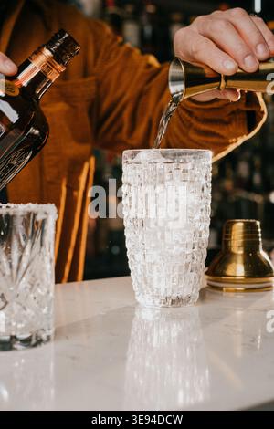 Un barista sta sta versando una bevanda in un bicchiere con ghiaccio. Il bicchiere si trova su un bancone accanto a una bottiglia Foto Stock