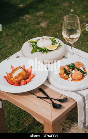 I piatti eleganti contengono piatti freschi e colorati. Un bicchiere di vino bianco aggiunge un fascino raffinato. Il tavolo all'aperto invita a una cena tranquilla e rilassata. Erba verde e natura Foto Stock