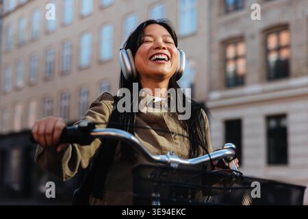 Una giovane donna ride mentre corre in bicicletta lungo una strada urbana, mette le mani sul manubrio e indossa cuffie sovrauricolari, ascoltando musica durante un su Foto Stock