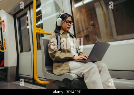 Un pendolare concentrato lavora su un notebook mentre è seduto in metropolitana, ascoltando le cuffie e concentrandosi su un'attività durante il tragitto in città, illustra Foto Stock