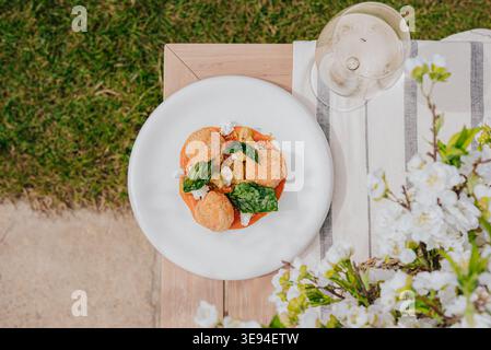 Il tavolo contiene un piatto elegante accanto al bicchiere di vino. L'erba verde incornicia i pasti all'aperto. I fiori bianchi aggiungono un fascino fresco e morbido. Piastre, vetro e tovagliolo Foto Stock