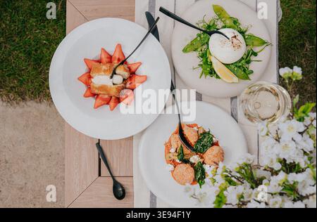 I piatti bianchi contengono piatti freschi e colorati. Fragole, verdure e cremosi condimenti rendono la scena invitante. Un bicchiere di vino seduto accanto al tavolo. Flusso Foto Stock