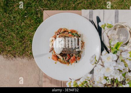 Pesce fresco su un vivace mix di verdure su un piatto bianco. Tavolo all'aperto con fiori e bicchieri accanto. L'erba e la superficie in legno incorniciano la ruggine Foto Stock
