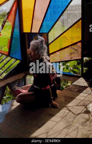 Donna matura seduta a gambe incrociate nel padiglione in vetro colorato di Bali Foto Stock