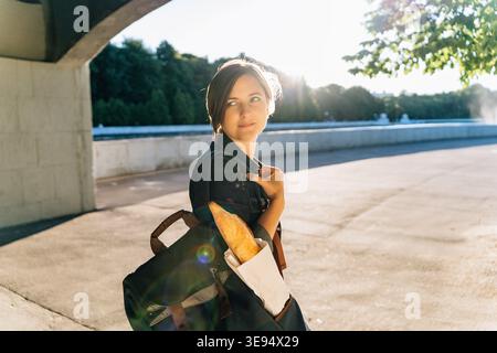 Attraente giovane donna con baguette nello zaino nel soleggiato parco cittadino Foto Stock