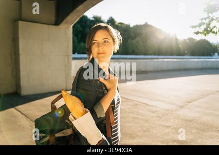 Donna contemplativa con baguette e zaino che cammina in una piazza del parco Foto Stock