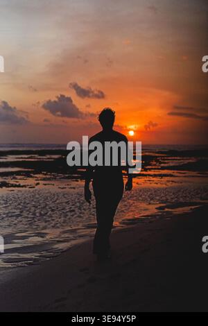 Silhouette di uomo che cammina sulla spiaggia al vivace tramonto arancione, Bali Foto Stock