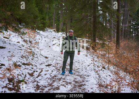 Un uomo maturo sorridente con zaino e macchina fotografica cammina verso la macchina fotografica lungo un sentiero nella foresta innevata attraverso un bosco di abeti rossi Foto Stock