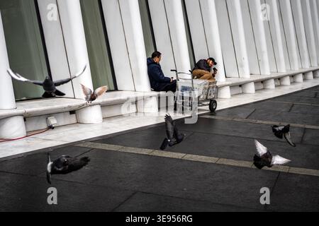 Una persona siede su una panchina mentre i piccioni volano e si aprono vicino a un edificio moderno con colonne bianche. Foto Stock