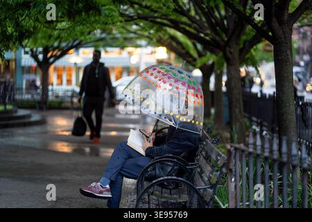 Una persona siede su una panchina del parco a leggere un libro sotto un ombrello colorato mentre cammina passa davanti a una persona che porta una borsa. Foto Stock