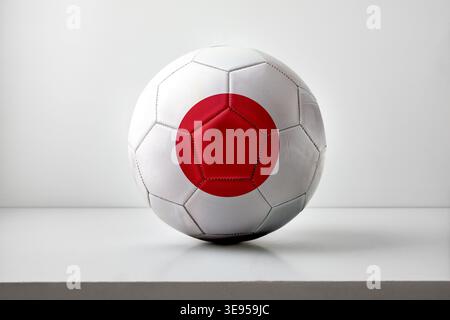 Un pallone da calcio bianco con il design della bandiera del cerchio rosso del Giappone, posizionato centralmente su un semplice sfondo bianco con un'illuminazione morbida e uniforme. Foto Stock