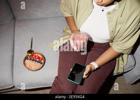 Donna adulta che tiene in mano smartphone e frullato durante una pausa salutare Foto Stock