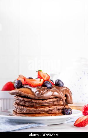 Pila di pancake al cioccolato con una ricca crema di pasta al cioccolato e frutti di bosco freschi - fragole, mirtilli. Colazione dolce e gustosa Foto Stock