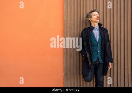 Uomo con capelli ricci appoggiato a un muro testurizzato, che guarda con un'espressione premurosa, contemplando nuove idee Foto Stock