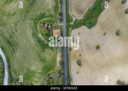 Vista aerea di una strada asfaltata rettilinea che divide prati verdi lussureggianti e campi arati secchi con un piccolo edificio in pietra a Pomarance, Toscana, Italia. Foto Stock