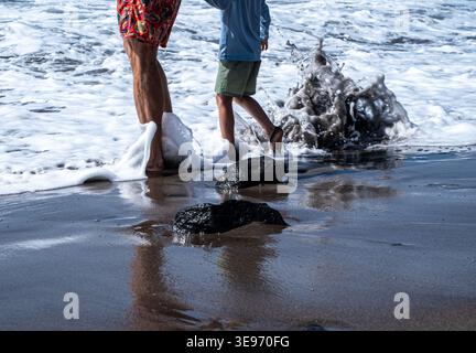 Un padre e un figlio camminano lungo la riva del mare. Le onde si infrangono intorno a loro mentre passeggiano lungo la riva. Foto Stock