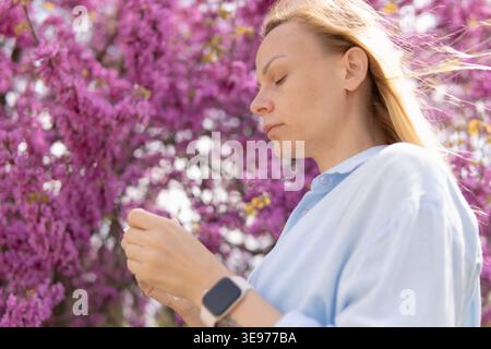Donna bionda con gli occhi chiusi, immersa nella calda luce del sole primaverile tra vibranti fiori rosa in un parco, godendo di calma, aria fresca e un momento di pace Foto Stock