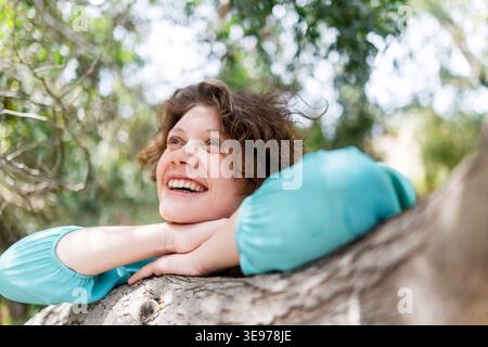Un ritratto ravvicinato di una donna che ride con ricci e lentiggini disordinati che appoggiano il mento su un tronco di albero. Foto Stock