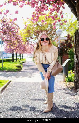Elegante donna in cappello che gode della stagione dei fiori di ciliegio, brillante ritratto all'aperto nel parco. Foto Stock