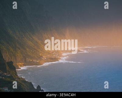 Vista aerea della costa frastagliata con ripide scogliere verdi e piccoli villaggi costieri durante l'ora d'oro a Madeira, Portogallo. Foto Stock