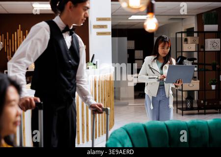 Giovane turista asiatico che cammina verso l'area salotto dell'hotel, tenendo in mano un computer portatile mentre è in vacanza. La donna che viaggia si reca in un punto wi-fi con un notebook nella lounge dell'accogliente resort Foto Stock