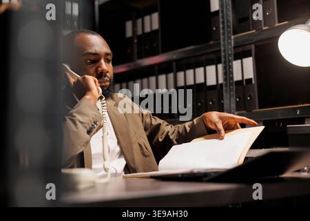 Investigatore della polizia afroamericana che risponde alle telefonate su rete fissa in ufficio di notte. Investigatore preoccupato che discute di informazioni sui casi di crimine mentre analizza il file del rapporto forense Foto Stock