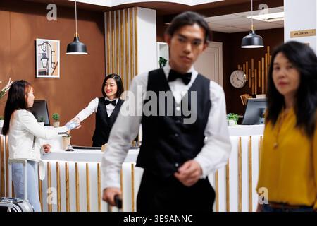 Asia Woman è guidata da un cordiale concierge che l'aiuta a trovare una camera, mentre un altro turista sta sta usando la carta di credito per pagare il suo soggiorno. Due ospiti dell'hotel si registrano nella hall Foto Stock