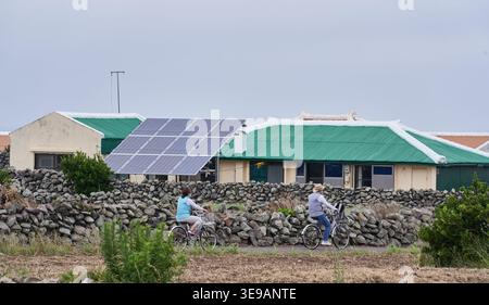 I ciclisti passano davanti a una casa rurale con un grande pannello solare. Questa vista panoramica mette in risalto la vita ecologica e le fonti di energia alternative. Foto Stock