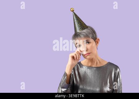 Triste donna matura in cappello da festa che piange su sfondo lilla Foto Stock
