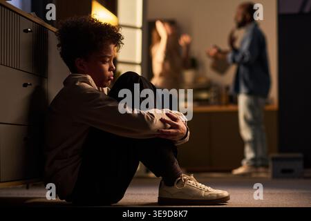 Adolescente seduto sul pavimento e abbracciato le ginocchia mentre i genitori discutevano in background, mostrando conflitti familiari e stress emotivo. Ideale per genitori, crisi domestica Foto Stock